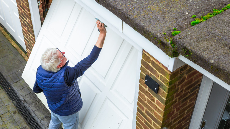 Beyaz garaj kapısının üstünü temizlerken mavi rüzgar kırıcı ve kazak giyen gri saçlı adam