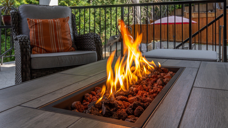 Gri hasır sandalye ve arka planda metal korkuluk bulunan bir masa merkezinde bir gaz ateşi.