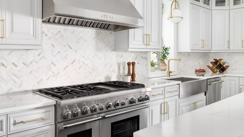 Gri dolaplar, balıksırtı karo backsplash, paslanmaz çelik önlük lavabosu ve altın donanım ve musluk içeren bir mutfak detayı.