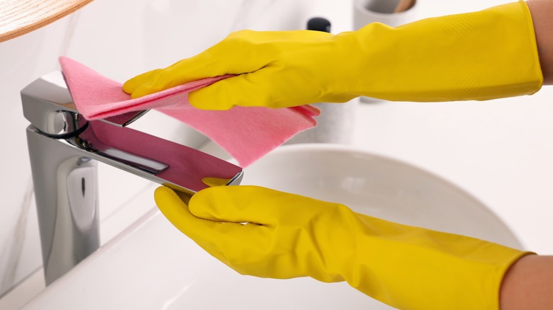 Bir banyo musluğunu silen eldivenlerde eller.