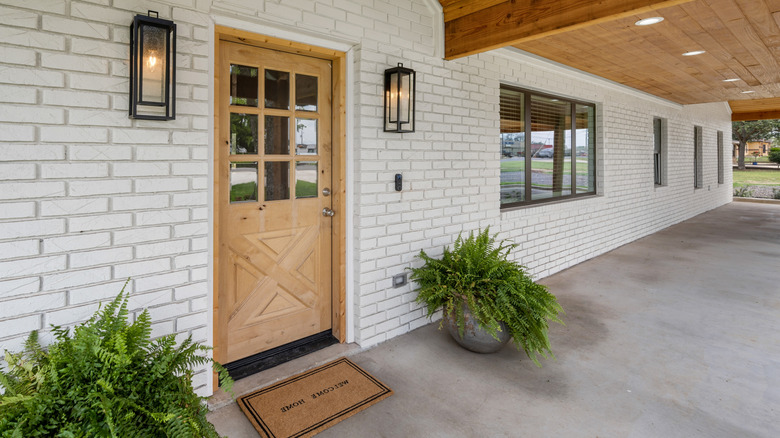 Ahşap ön kapılı beyaz bir tuğla ev, aydınlatma aplikleri ve eğrelti otları