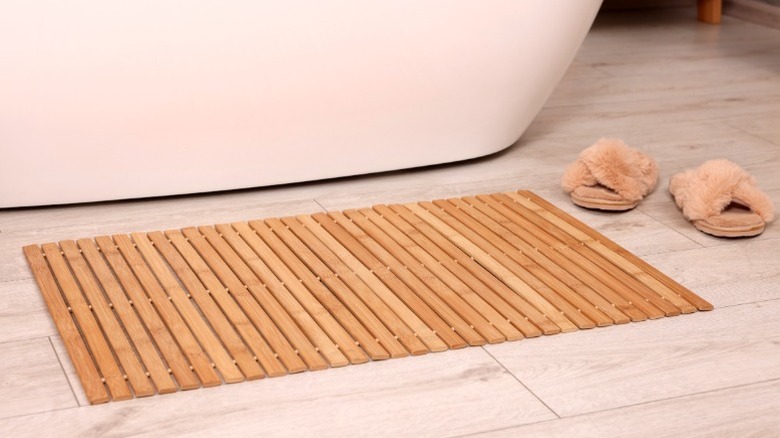 Terlik ve beyaz küvetin yanındaki fayans banyo zeminde oturan bambu banyo mat