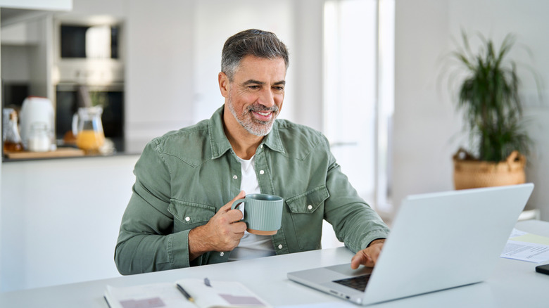 Kahve kupasından içerken dizüstü bilgisayarda kaydıran yaşlı adam