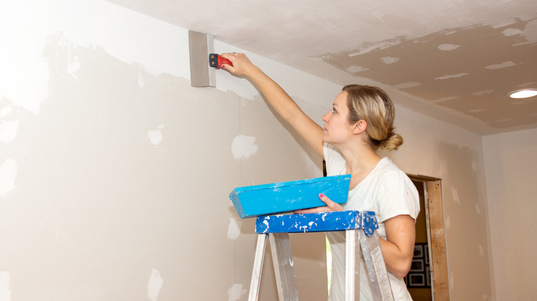 Bir kadın, yeni asılı alçıpandaki eklem bileşiğini yumuşatırken bir merdiven üzerinde durur