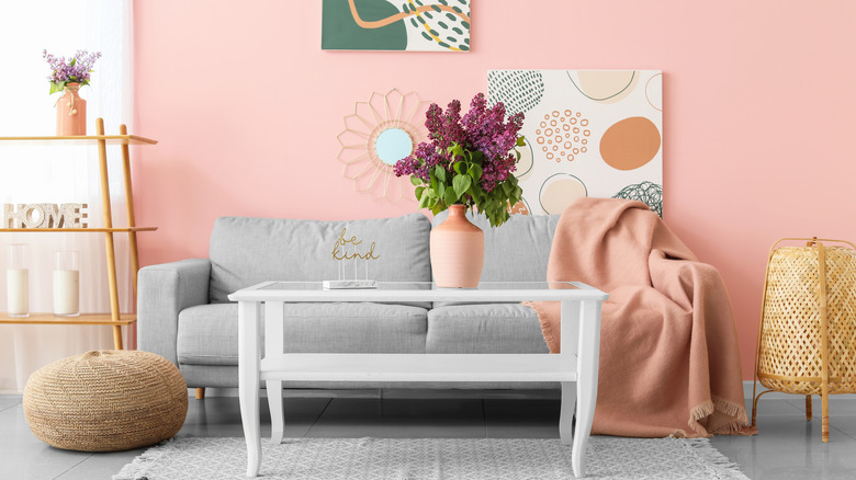 Duvarda sanat eseri olan pembe bir oda, beyaz bir sehpa, gri bir kanepeye örtülmüş bir mercan battaniyesi, raflar ve hasır dekor