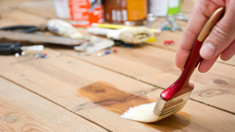 Bir adamın eli leke veya dolgu macunu kaplı bir fırça tutar ve bir ahşap güvertenin tahtasına aktarır.