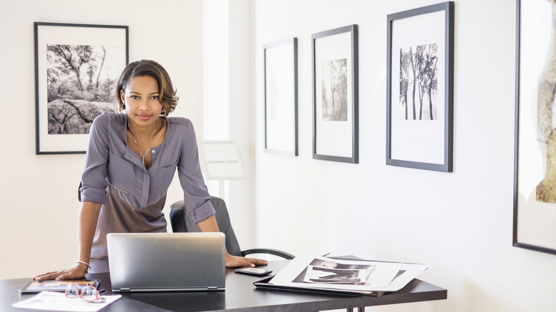 Bir kadın, bir dizüstü bilgisayar ve diğer eşyaları olan bir masanın önünde durur, çünkü arkasındaki duvarlara farklı çerçeveli portreler asılır.