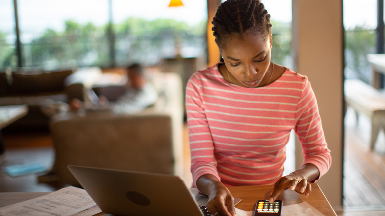 Dizüstü bilgisayarının önünde bir masada oturan kadın, hesap makinesi uygulaması telefonda açık