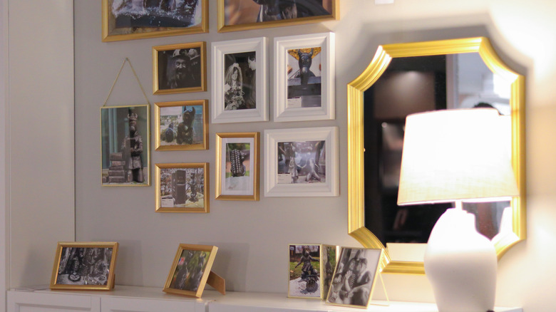 Altın ve beyaz çerçevelerde çeşitli çerçeveli fotoğraflara sahip bir galeri duvarı, dekoratif bir ayna ve modern bir lamba içeren şık bir iç ekran.