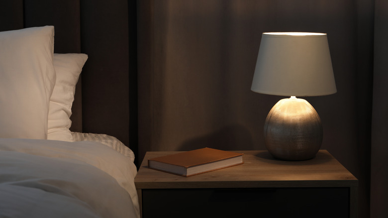 Bir kitap ve yanındaki yatak ile ahşap bir komodin üzerinde hafif bir başucu lambası.