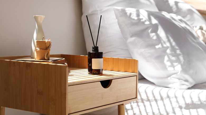 Difüzör, vazo, mum ve yatak köşesi ve arkasında beyaz yastıklarla kitap yığını ile başucu masası.