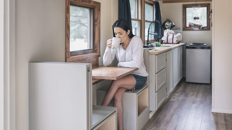 Kadın, küçük bir evde ahşap bir kabinde bir kupadan içiyor oturuyor