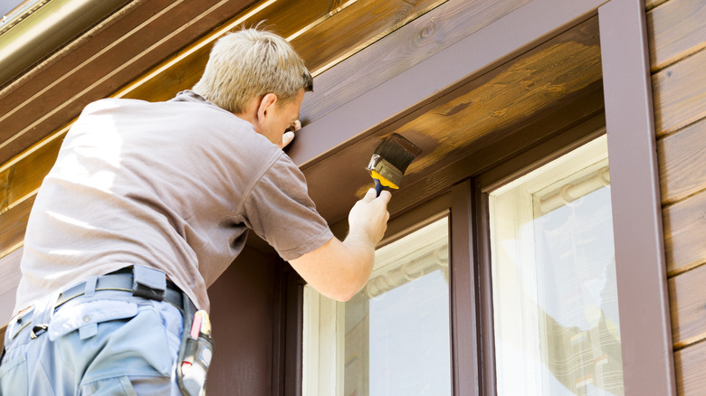 Pencerenin üzerindeki ahşap bir dış duvara fırça ile kahverengi boya uygulayan işçi.