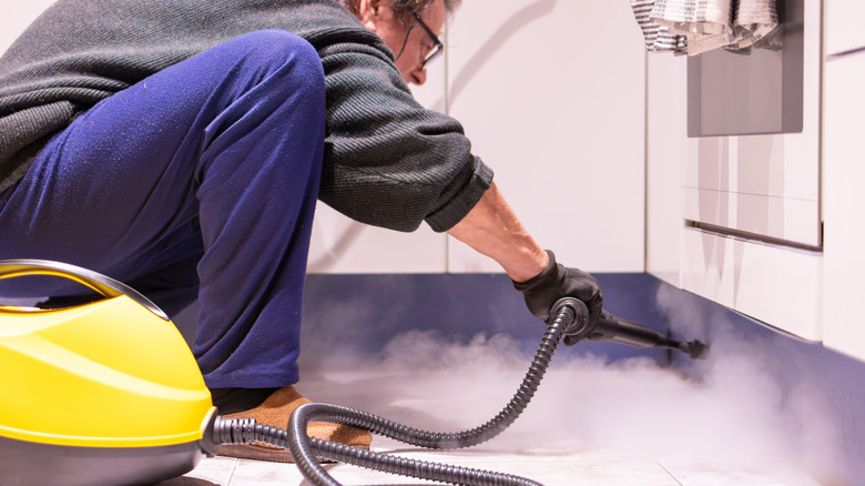 Bir adam, cihazların altındaki mutfak duvarlarını temizlemek için bir vapur kullanarak bir mutfak tabanına çömelir.