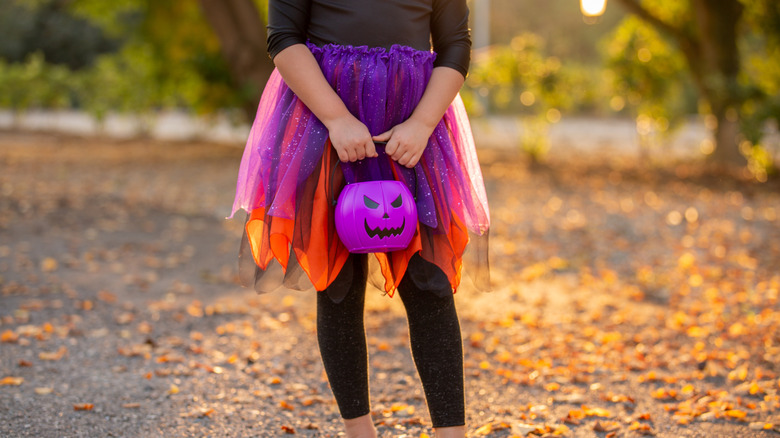 Düşen sarı yaprakların olduğu bir sokakta mor bir Cadılar Bayramı kovası tutan çocuk