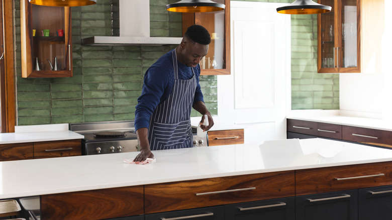 Adam mutfakta beyaz kuvars tezgahı ahşap dolaplarla siliyor.