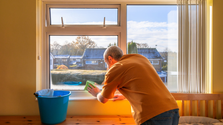 Yakınlarda mavi bir kova dururken süngerle pencereyi temizleyen bir adam