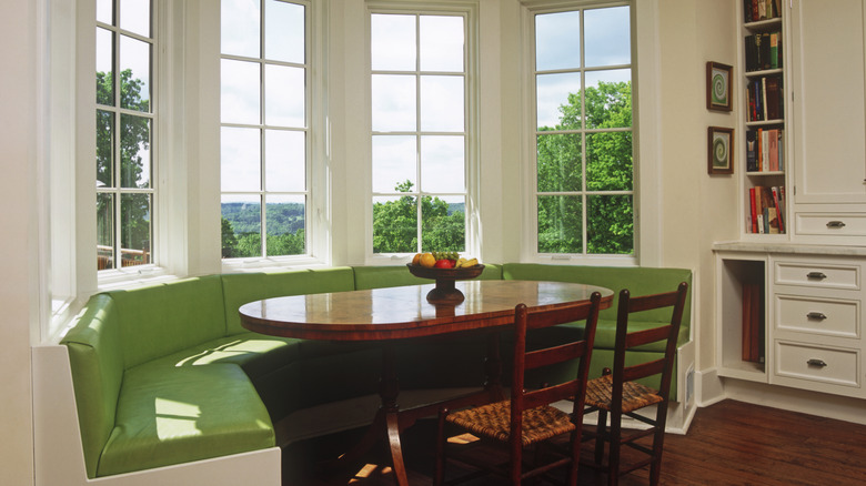 Cumbalı pencerenin önünde oval masa ve iki hasır sandalyeden oluşan yeşil banket oturma alanı.