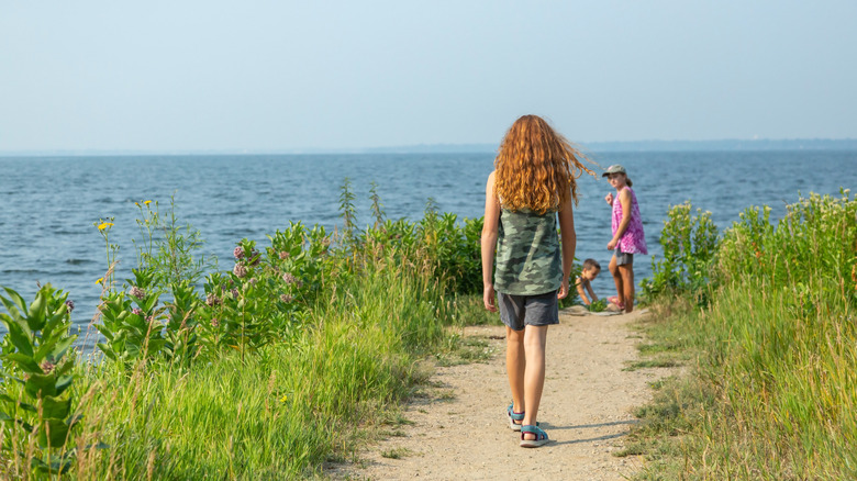 Kızıl saçlı bir kız, arka planda aile üyeleriyle birlikte göl kıyısındaki bir yolda yürüyor.
