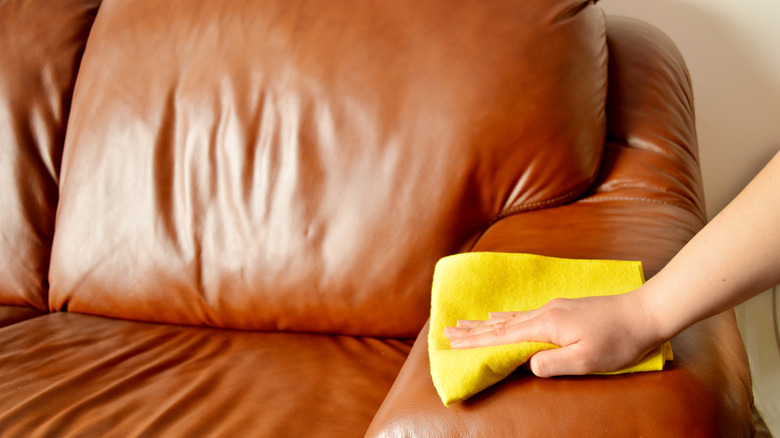 Bir kişi deri kanepeyi sarı mikrofiber havlu kullanarak temizliyor.