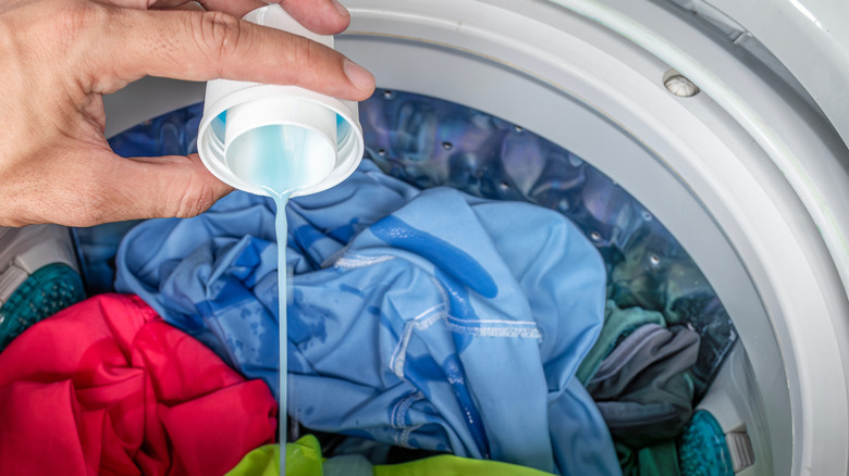 Bir kişi çamaşırlarla dolu çamaşır makinesine çamaşır deterjanı döküyor.