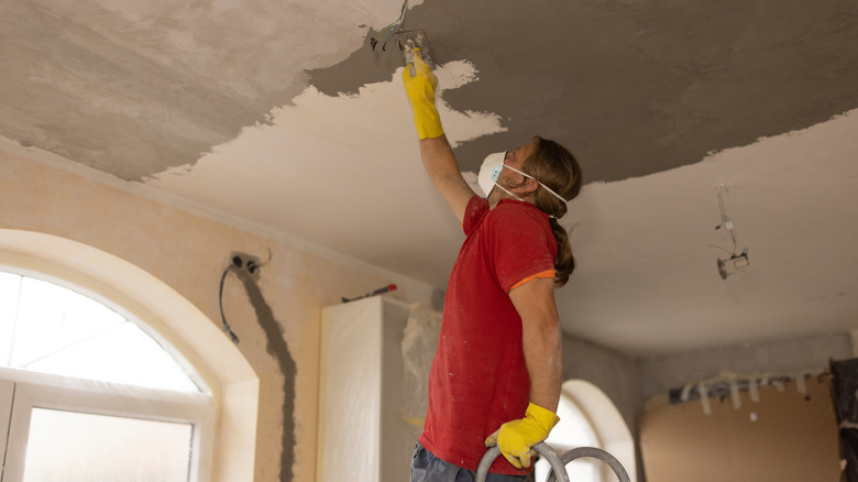 Maske takan bir adam merdivende dururken tavana macun sürüyor