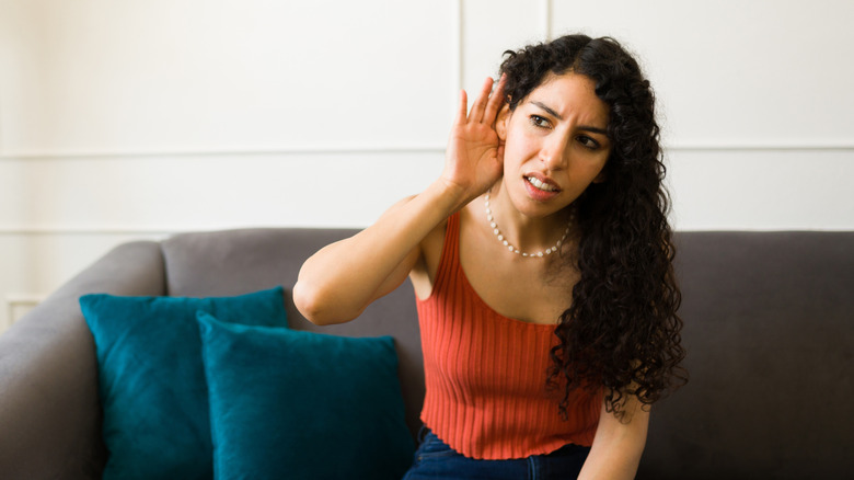 Kanepede oturan bir kadın sesi dinlemek için elini kulağının arkasına götürüyor.