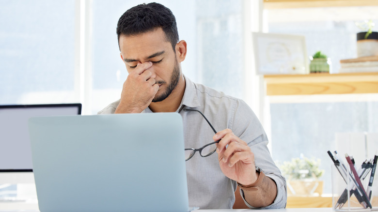 Başı ağrıyan bir adam bir eliyle gözlüğünü tutuyor, diğer eliyle burun köprüsünü sıkıştırıyor.