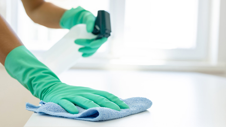Yeşil eldivenli biri beyaz masanın yüzeyini sprey şişesi ve bezle temizlemeye başlıyor.