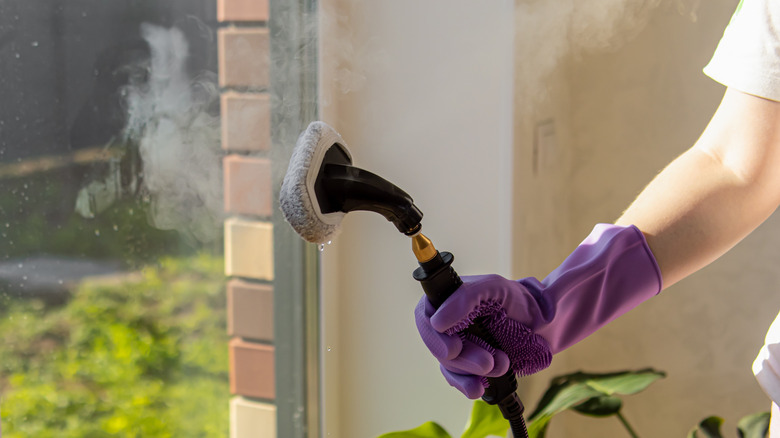 Mor eldiven giyen bir kişi buharlı temizleyiciyi pencereye doğru tutuyor.