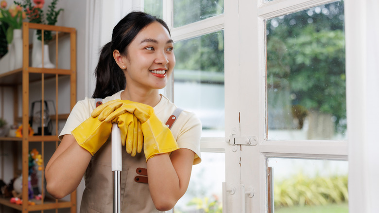 Eldiven giyen bir kadın paspas sapına yaslanarak temiz bir pencereden dışarı bakıyor.
