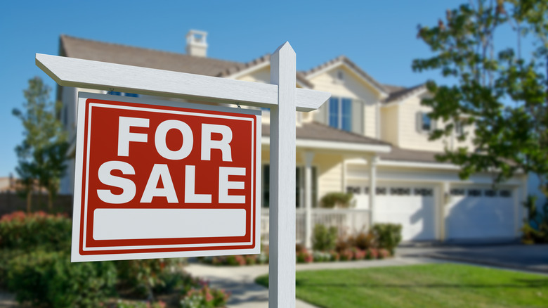 Bahçesinde kırmızı ve beyaz SATILIK tabelası olan satılık ev, sarı bir evin bulanık odağı