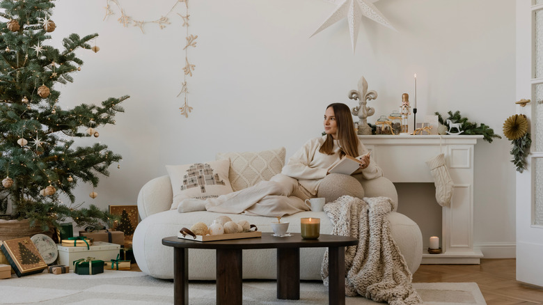 Gülümseyen kahverengi saçlı bir kadın, Noel için dekore edilmiş nötr renkli bir odada kitap okuyor.