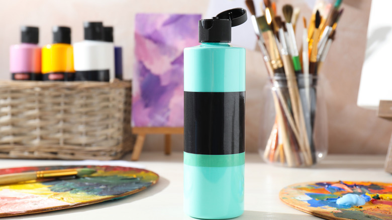 Arka planda diğer boyama malzemeleriyle birlikte açık bir akrilik boya şişesi masanın üzerinde duruyor.