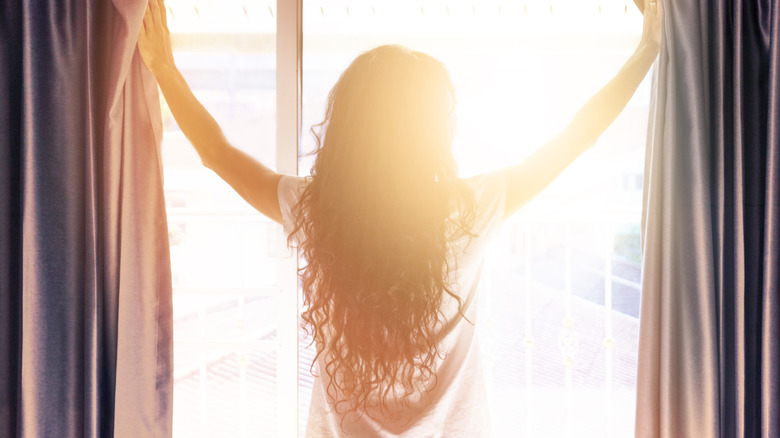 Kadın perdeleri açıyor ve güneşi içeri alıyor.