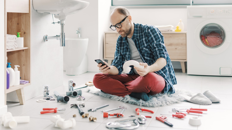 banyo zemininde oturan ve etrafı aletlerle çevriliyken telefonu kaydıran adam