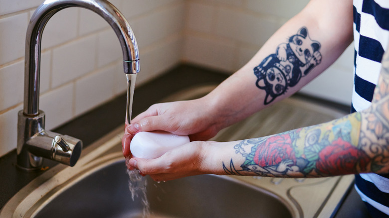 Dövmeli kişi mutfak lavabosunun altındaki sabunu duruluyor.