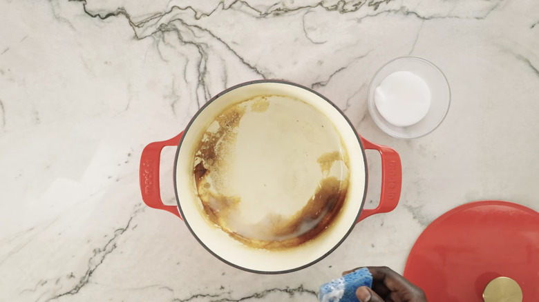 Bir kişi kırmızı emaye dökme demir tencereyi sirke, fırın kanepesi ve mavi süngerle temizler.