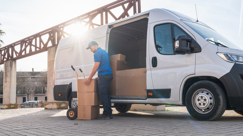 Teslimat sürücüsü, el kamyonuyla paketleri minibüsten boşaltıyor. Bir minibüste teslimat sürücüsü konsepti. Mavi üniformalı adam, teslimat aracından kutuları boşaltmak için el arabası kullanıyor