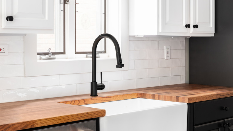 Beyaz çiftlik evi lavabosu, siyah musluğu ve beyaz backsplash'ı olan koyu renkli kasap blok tezgahı.