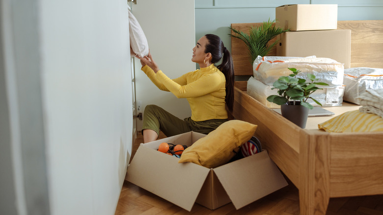 Bir kadın, kutular ve çarşaflarla çevrili bir yatak odasında eşyaları ayırıyor ve düzenliyor.