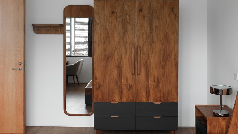 Yanında bir ayna bulunan, alt kısmı siyah raflı, duvara dayalı ahşap bir gardırop