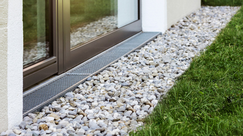 Bir tarafında sürgülü cam kapı, diğer tarafında yeşil çim bulunan bir evin yakınındaki kanalizasyon yolu