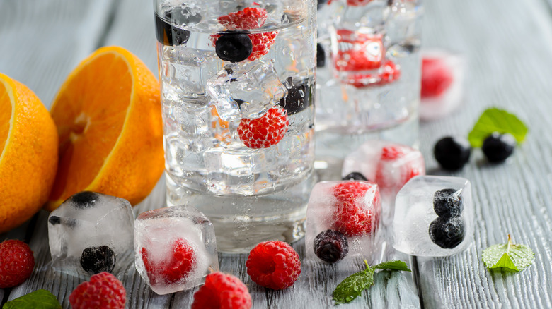 Meyvelerle ve buz küplerinde donmuş meyvelerle çevrili ahşap bir masanın üzerinde su, buz ve meyvelerle dolu bir cam bardak