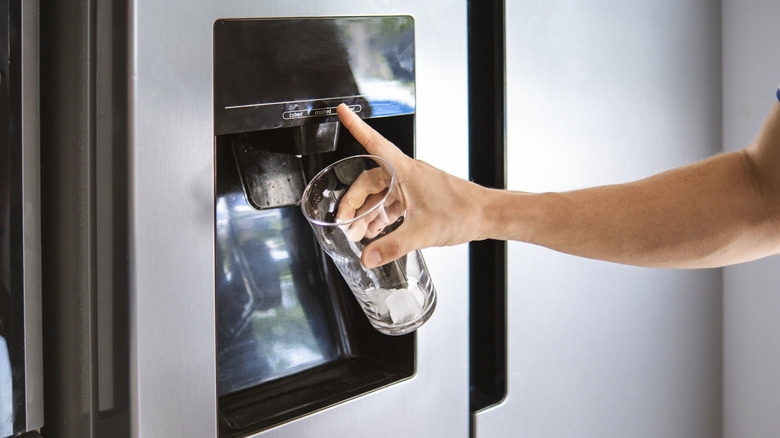 Elinde birkaç buz küpü olan cam bir bardakla buzdolabı buz makinesinin ve su pınarının düğmelerine basan bir el.