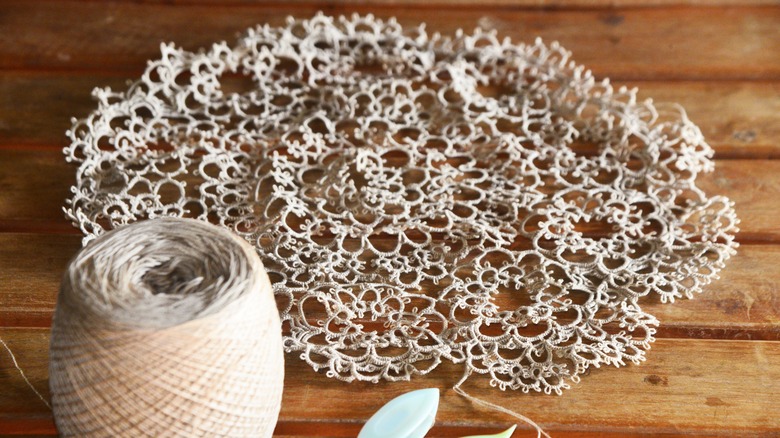 Ahşap, çıtalı bir yüzey üzerinde bir makaranın yanında duran yuvarlak bir dövme dantel parçası.