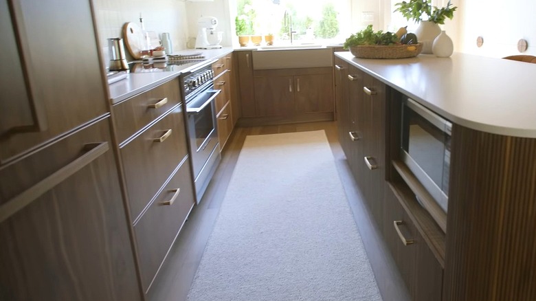 Ceviz ağacından ahşap dolapları ve nervürlü dış cephe kaplaması ve beyaz tezgahı olan ceviz ağacından adanın bulunduğu mutfağın görünümü. Adanın içindeki bir rafa bir mikrodalga fırın yerleştirildi.