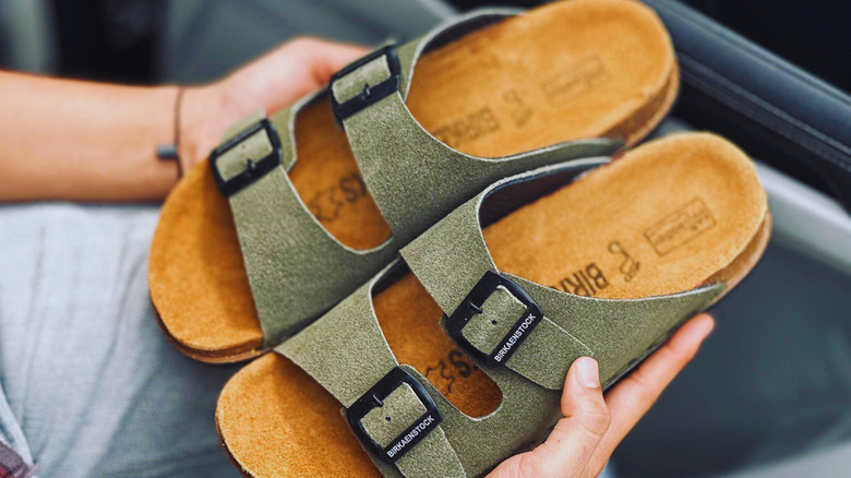 Bir çift yeşil Birkenstock sandaleti tutan bir adam.