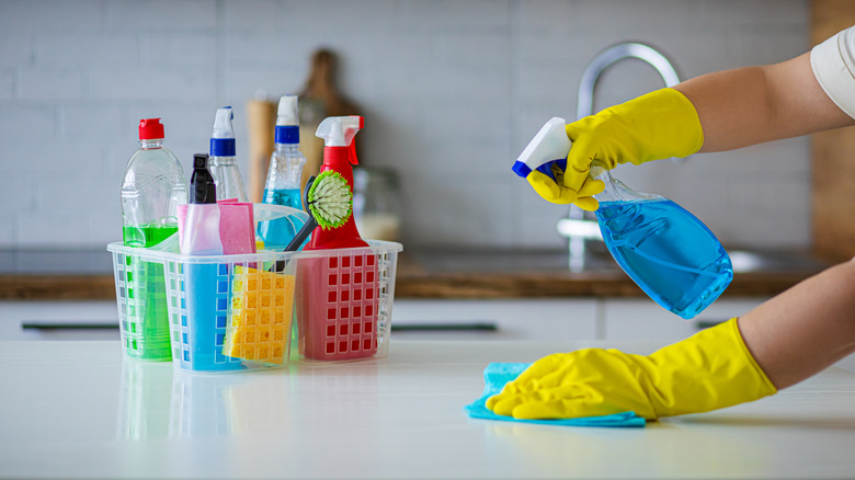 Dolu temizlik kutusunun yanındaki tezgahı spreyleyen ve silen kişi