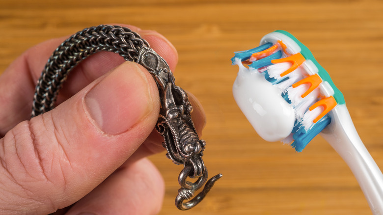 Bir elinde diş macunu olan bir diş fırçası, diğer elinde kararmış bir gümüş takı parçası tutan bir kişi var.